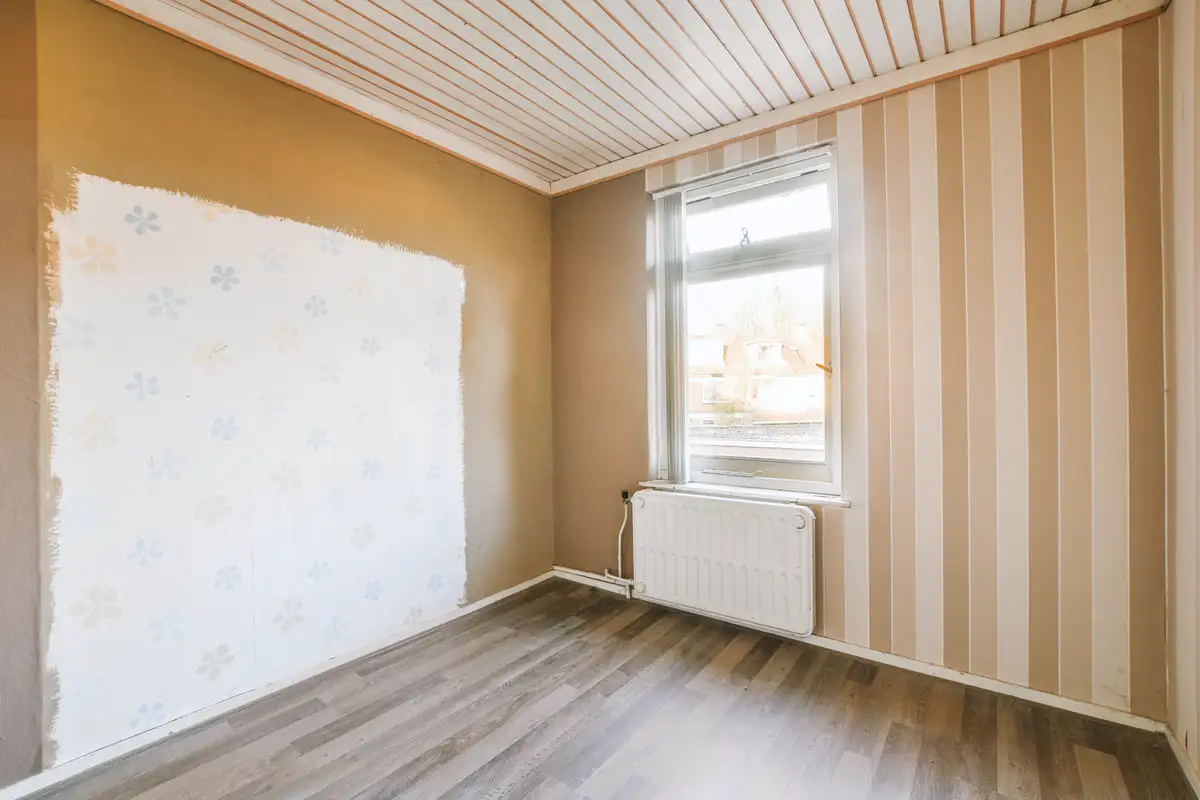 Leerer Raum mit teils abgezogener Wandtapete, Heizkörper unter Fenster und Laminatboden – Vorbereitung für Tapezieren